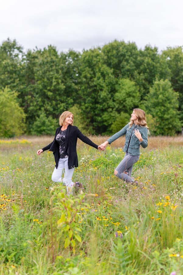 Two women holding hands and smiling, joyfully spinning in a lush meadow filled with wildflowers.