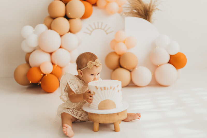 Cake smash session: A little girl playfully bites into a cake amidst vibrant balloons in a professional photo studio.