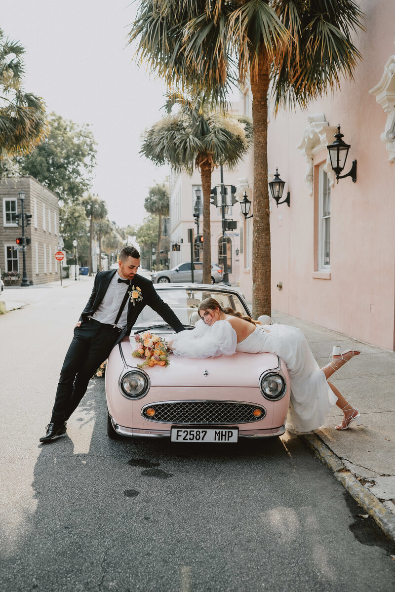 A man in a suit and a woman in a wedding dress pose leaning on a pink vintage car on a city street lined with buildings and palm trees.