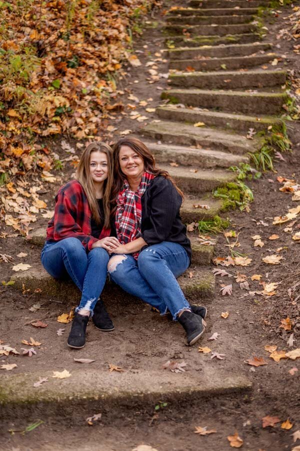 Two women wearing plaid shirts and jeans sitting together on wooded stair steps, surrounded by autumn leaves.
