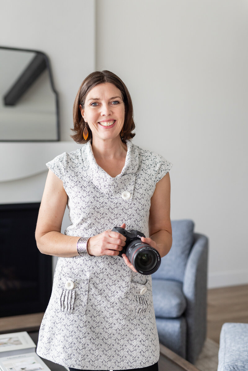 Calgary creative branding photographer, Kate Daniel Photography, standing and holding her black camera while wearing a white, patterned tunic with yellow, leaf earrings.