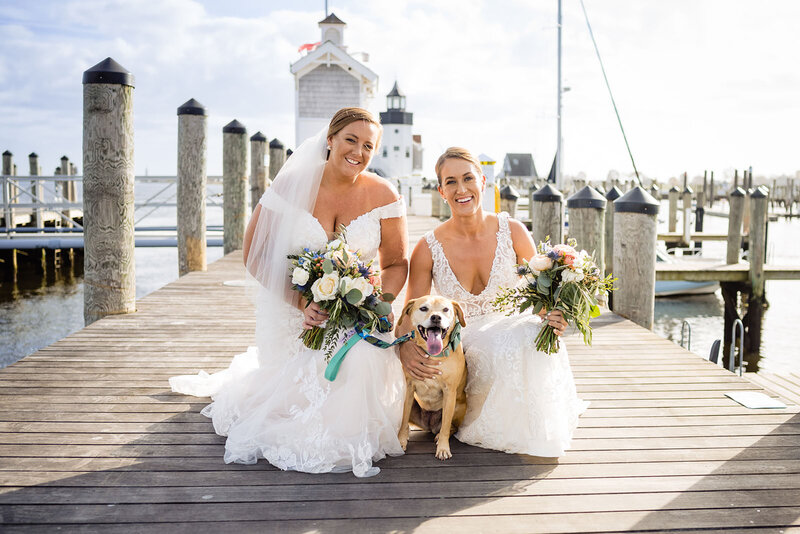 Two brides squatting down with a dog on a pier