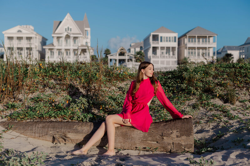 A sixteen year old girl sits on a driftwood log on Galveston beach in front of dune and beach houses