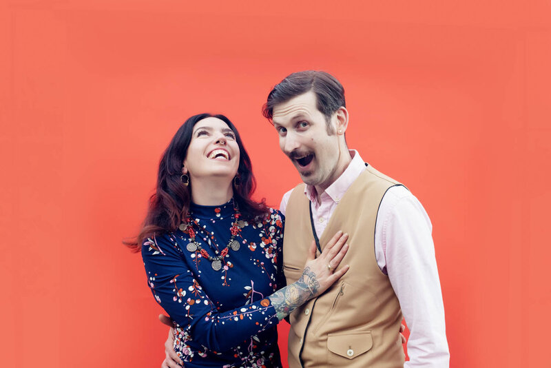 a silly and fun couple pose for the camera  in front of a bright orange wall during their engagement photos