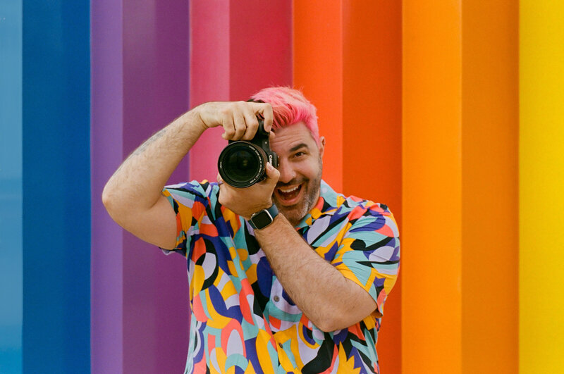 A photographer holding a camera up to their head in front of a rainbow wall.