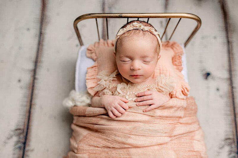 A sleeping newborn baby is wearing a peach outfit laying in a bed with a peach blanket