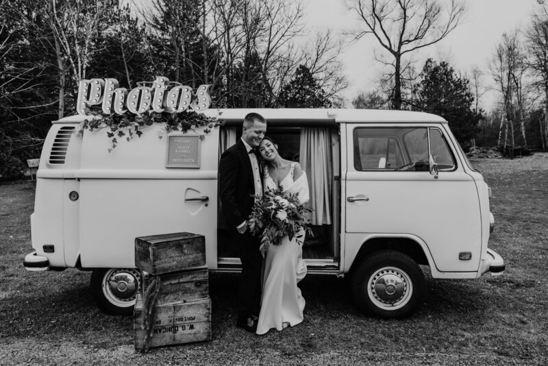 Bride and groom pose for a fun couples wedding portrait photo at Holland Marsh Wineries in Newmarket, ON. Black and white image of bride and groom cuddling and laughing in front of a vintage VW camper van that was converted into a photo booth. Greenery and a lighted "photo" sign decorate the VW camper van for this wedding.