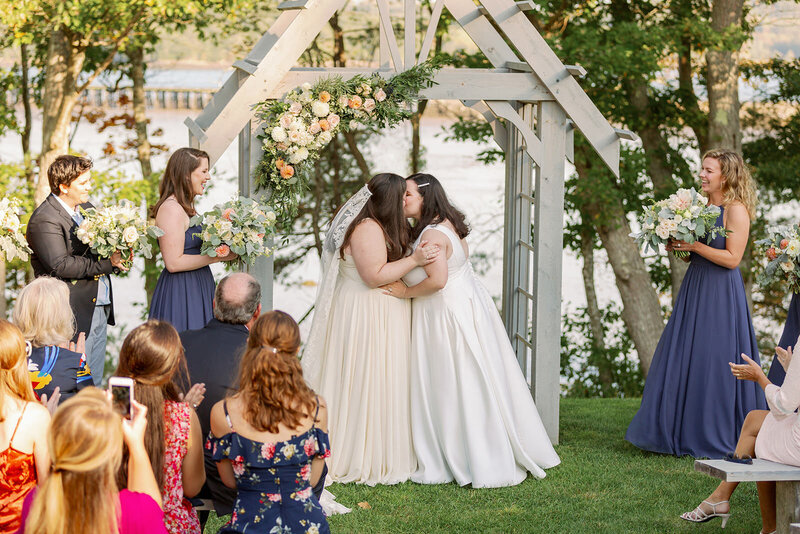 Two brides during their first kiss with their wedding parties and guests clapping.