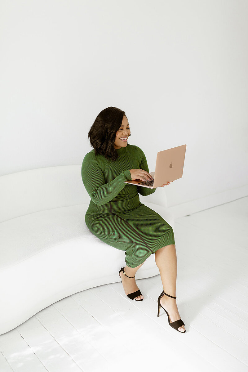Businesswoman in a green dress sitting on a white sofa, working on a gold-colored laptop.