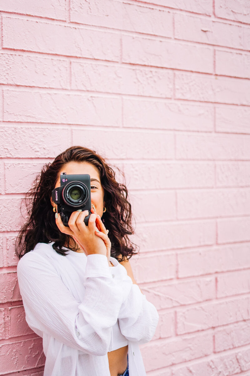 Gen Z woman wearing white t-shirt in front of pink brick wall aims her camera at the person taking her picture