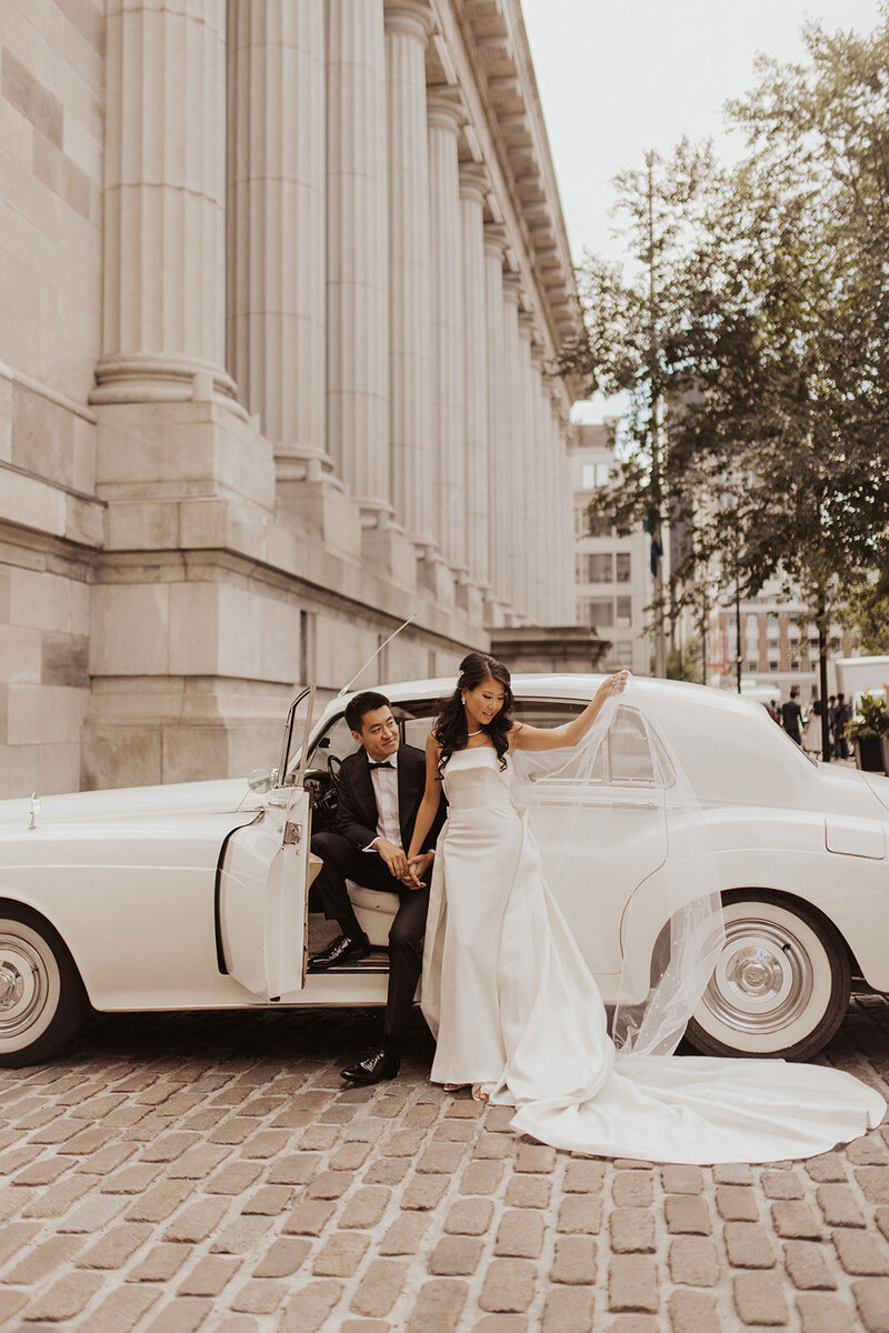 Couple by vintage car, bride adjusting veil, urban wedding shoot.