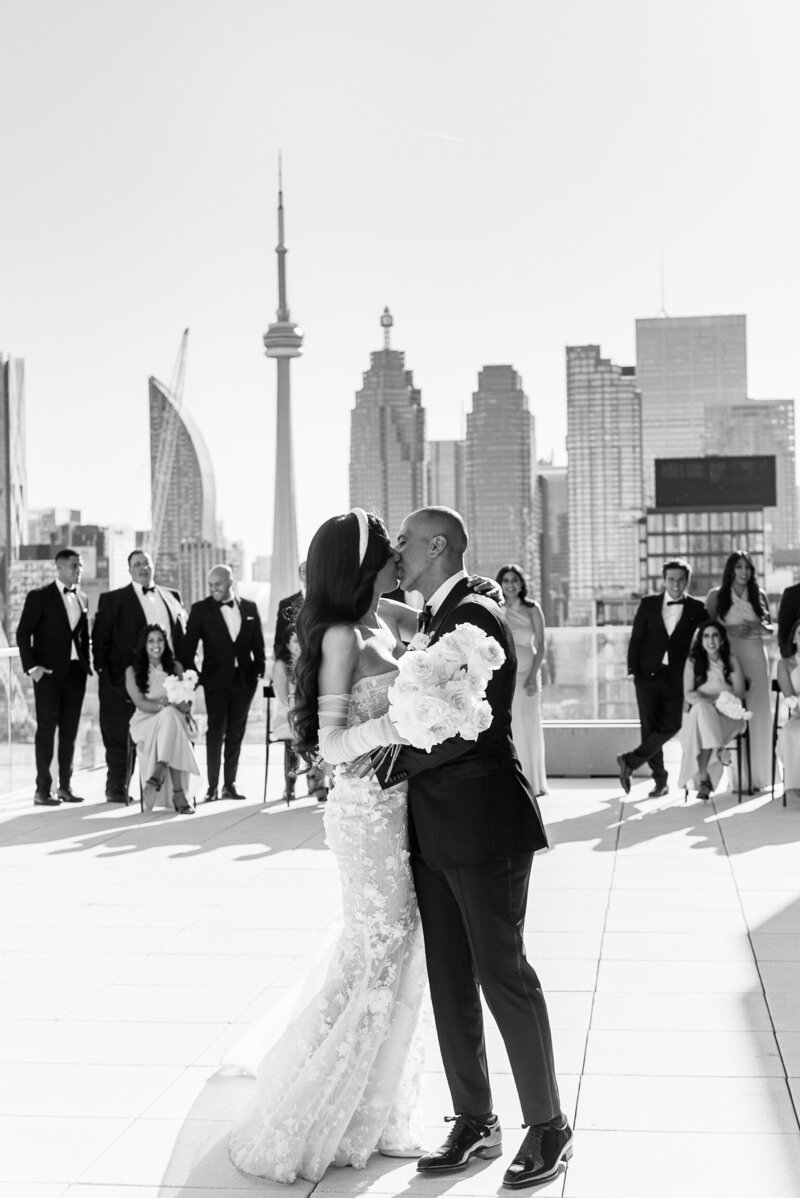 Wedding couple kissing on a rooftop with their bridal party and Toronto skyline behind them