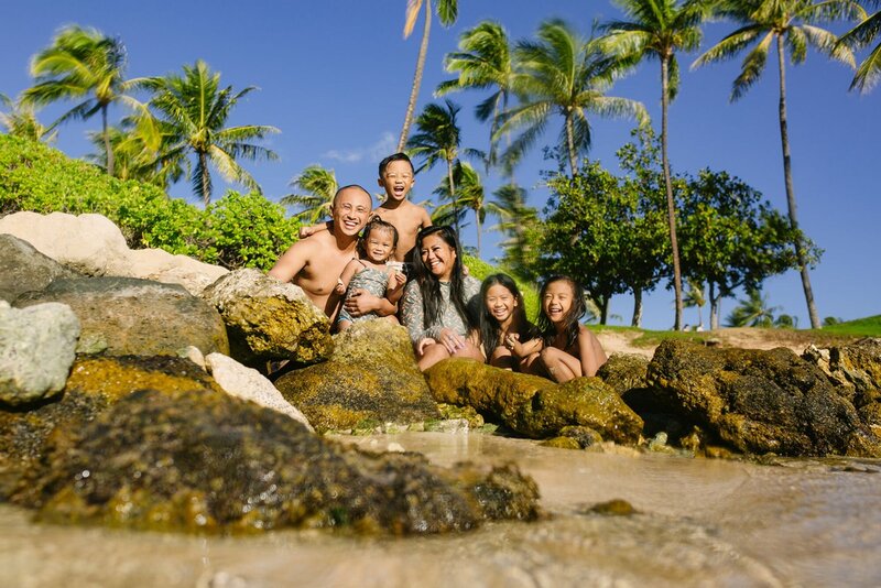 A family with four children laugh and smile as they sit on rocks in a lagoon.