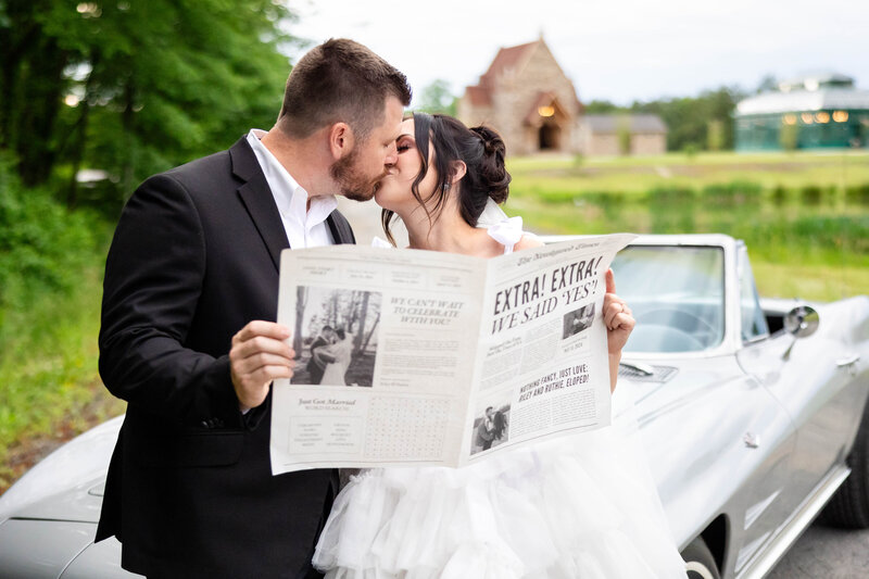 Bride and Groom sitting on the front of their classic car holding up a newpaper that announced their wedding.  Bride in a white short ruffled flirty dress and groom in a black tuxedo.  The couple are kissing.  The venue of a chapel and atrium can be seen in the background.