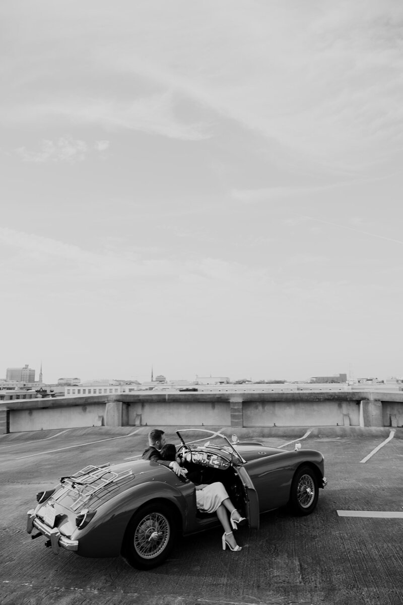 Black and white photo of classic sports car on top of parking garage. Couple sitting inside. Woman has legs out of open door. CHarleston city in background