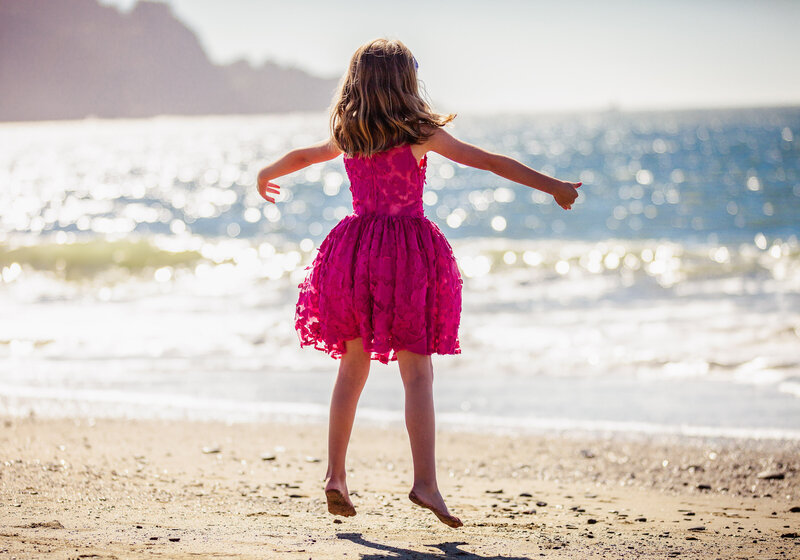 Girl in pink dress does ballet jump on Baker Beach in San Francisco, CA