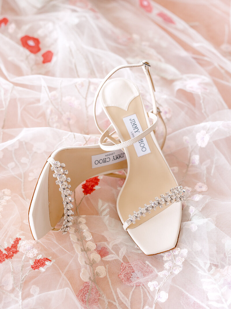 Jimmy Choo wedding shoes laying on top of a veil with red flowers on it