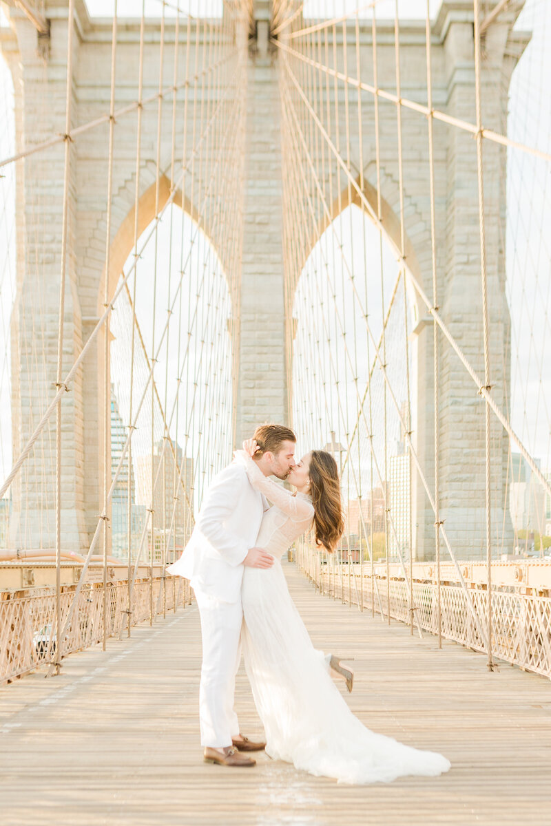 Bride and groom stand on a steel truss pedestrian bridge with the NYC skyline in the background. The bride and groom are facing each other and share a fun kiss with the bride's foot kicked out behind her. Captured by top NYC Destination Wedding Photographer Lia Rose Weddings