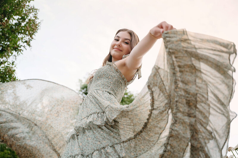 Senior photos of a girl twirling around outside in a floral dress, smiling, and looking down at the camera
