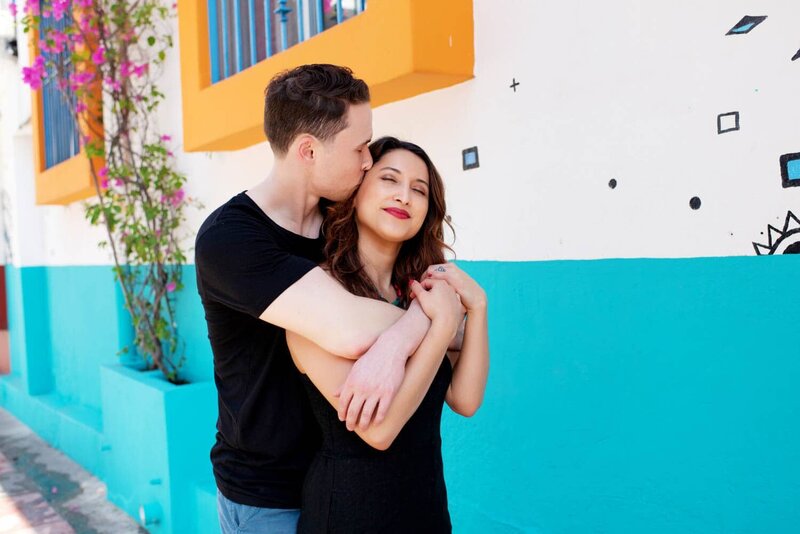an engaged man and woman standing in front of a very colorful and fun blue and orange wall in puerto vallarta hug each other