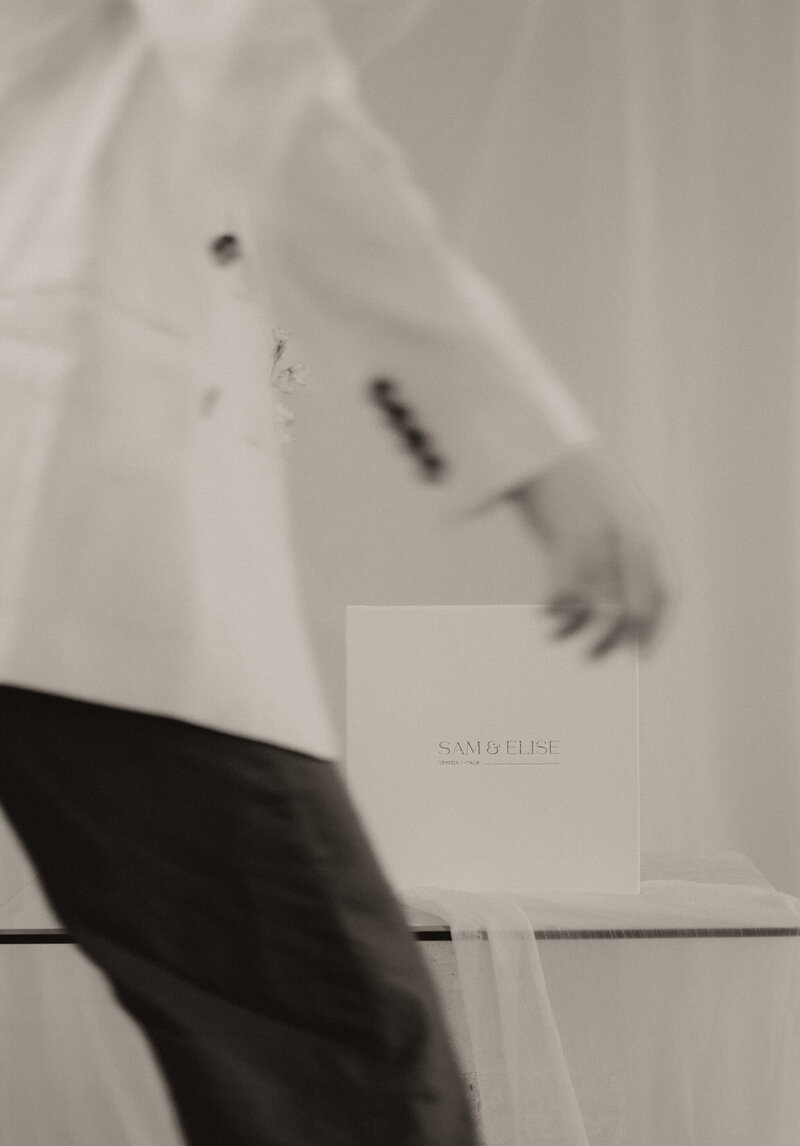 Black and white photo of man walking in front of  the camera that is focused on a white wedding album set on a  table- Romero Album Design