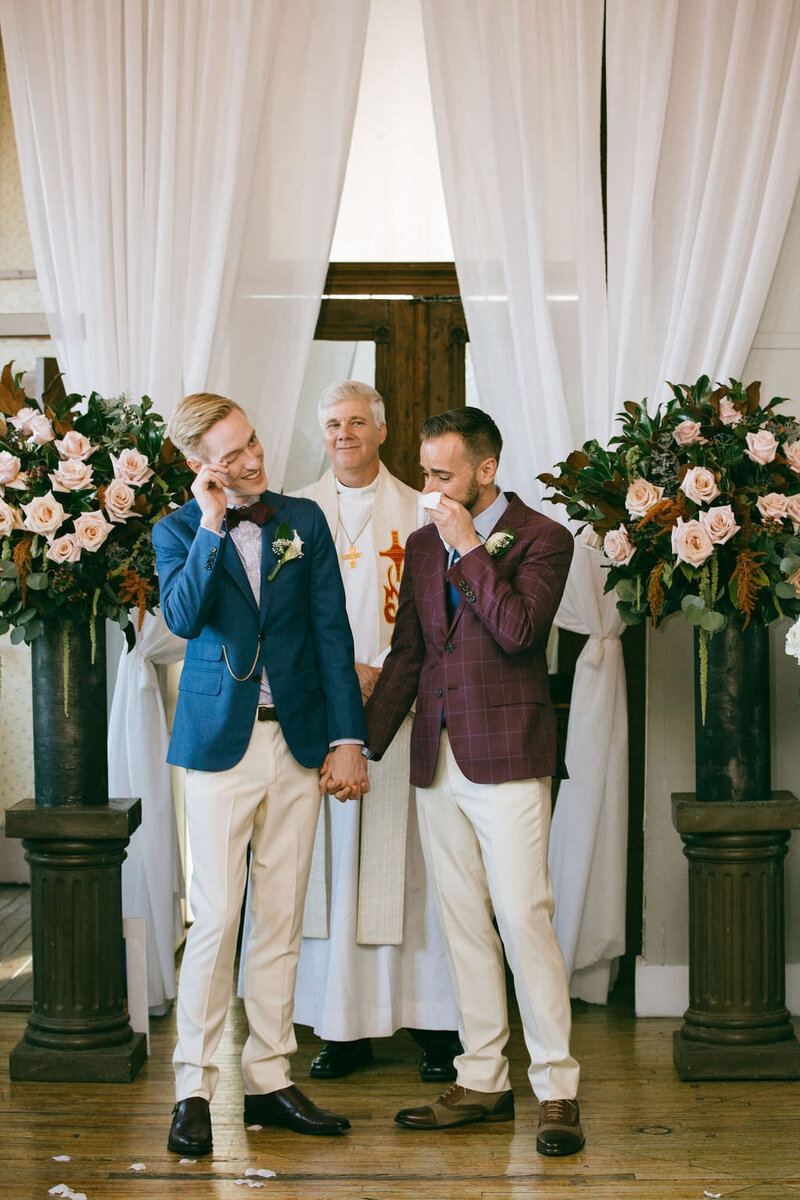 Two grooms holding hands and crying during their wedding ceremony.