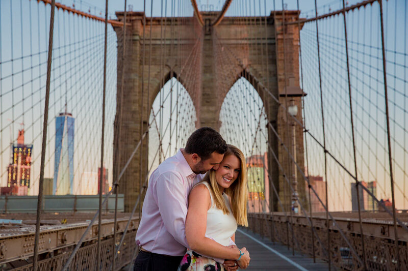 A man with his arms around a woman as he leans down to kiss her cheek on a large bridge.