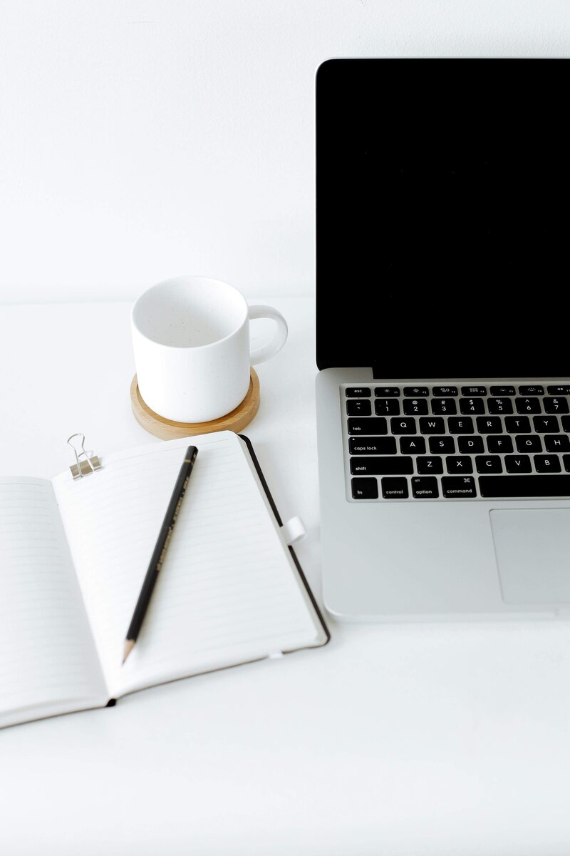 A clean, minimal workspace featuring a closed notebook with a pencil on top, a white mug on a wooden coaster, and an open laptop with a black screen on a white desk.