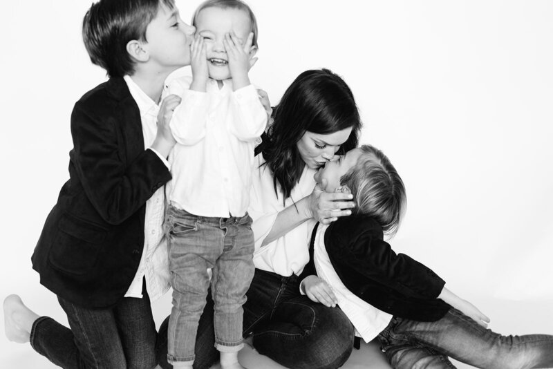 A mother kisses her young child while her oldest child kisses his younger brother on the cheek.