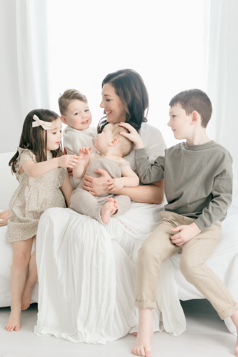 Mom with 4 of her young children all smiling at one another in all white studio in NJ