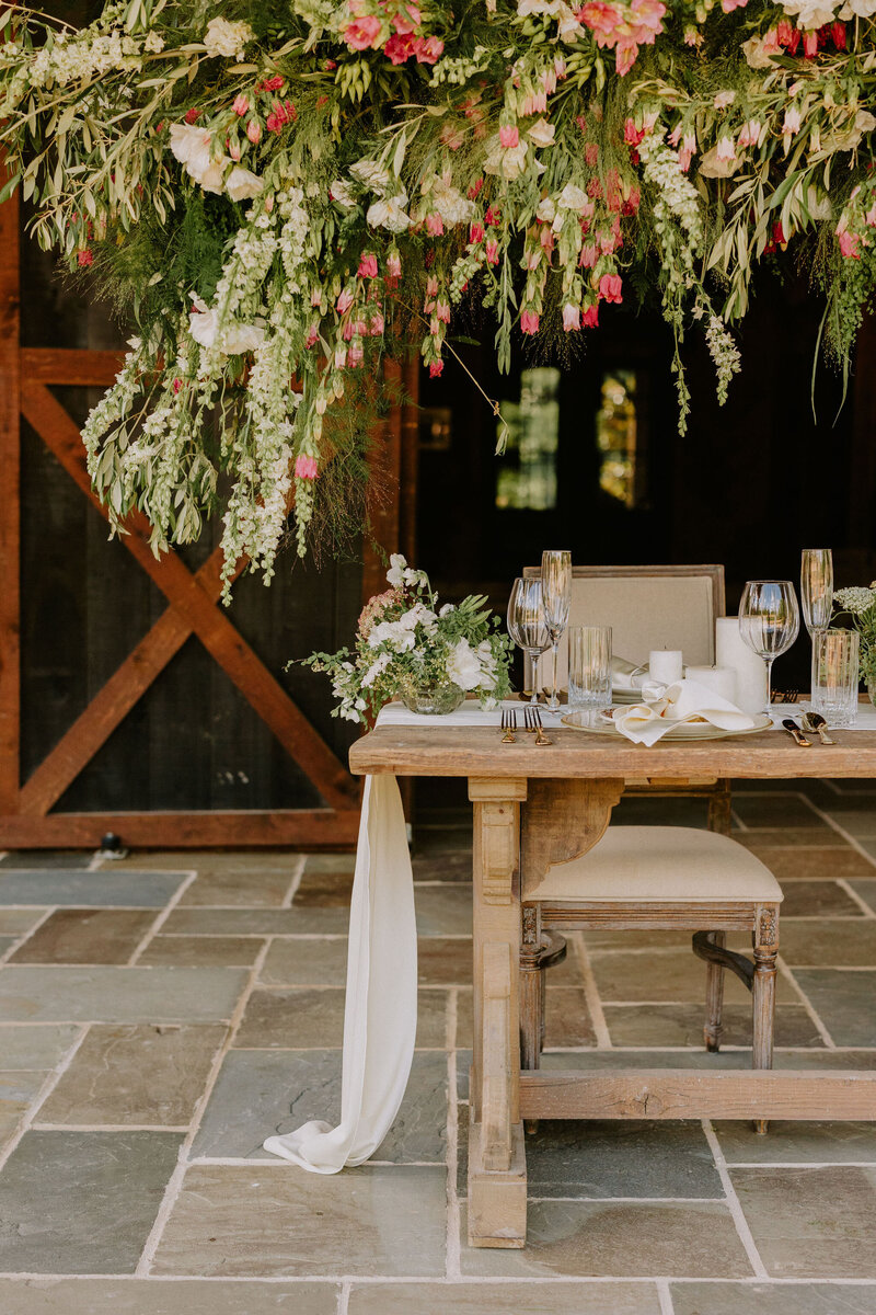 The left side of a wooden reception table with a suspended floral design above it and centerpieces on top of the table at a Nashville wedding