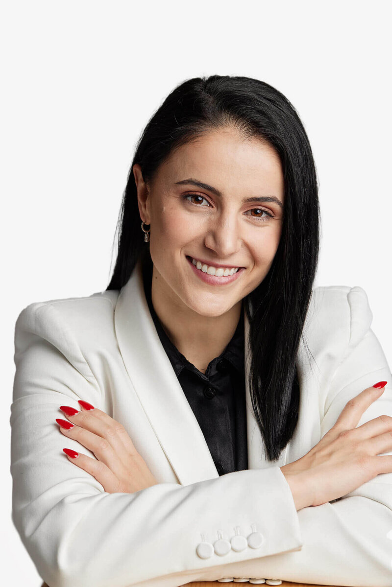 Woman in white jacket and black shirt against a grey background. She's smiling and has red nail polish.