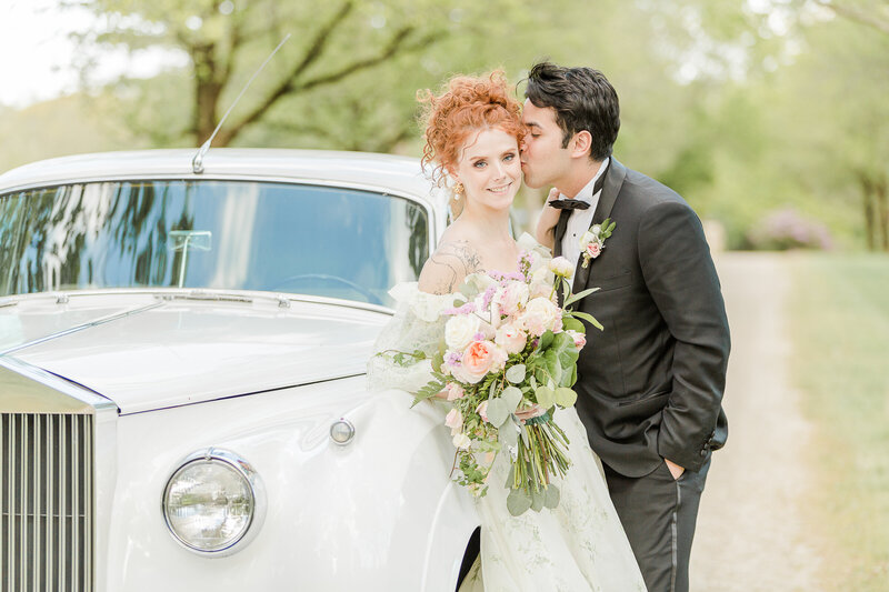 Bride with upswept curly red hair is leaning against a white Rolls Royce for a wedding day portrait. Her groom in a tuxedo is gently kissing her cheek. The bride is smiling at the camera and is holding her flowers in front of her.