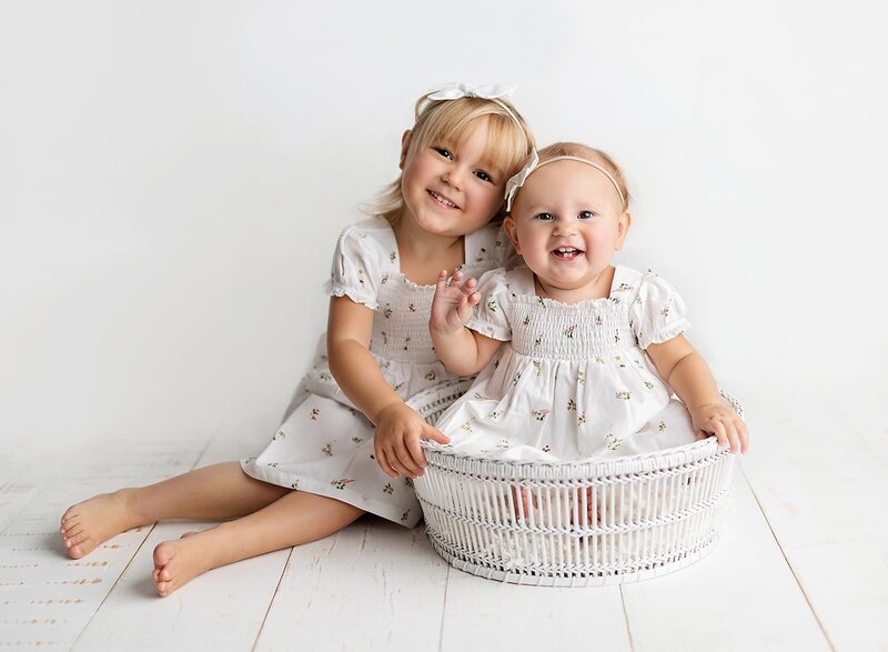 A toddler girl sits on a studio floor with her little sister in a matching dress in a basket posed by a Lafayette Baby Milestone Photographer