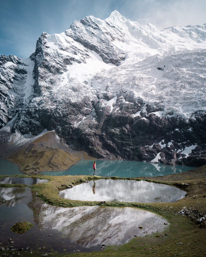 Woman hiking standing next to a pool of water and a large snow covered mountain in Peru