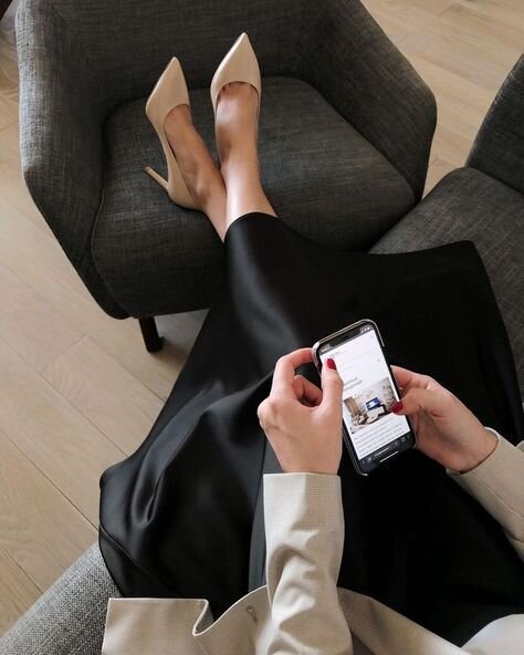 Woman in black skirt and tan heels holding a cell phone with feet propped up on a grey chair