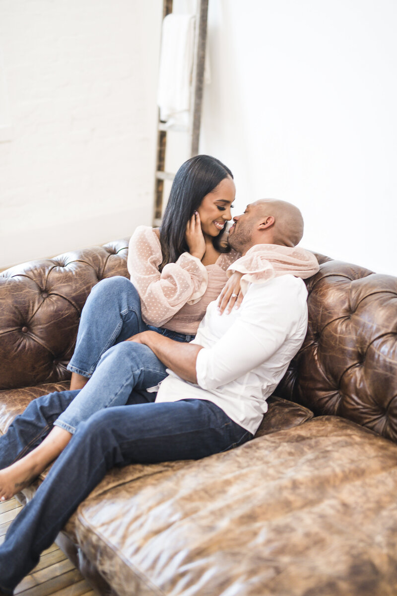 A couple cuddles on a rustic brown leather couch in an industrial photography studio.