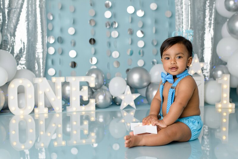 A one year old little boy sits in a blue diaper cover, suspenders, and a boy tie during his studio cake smash session.