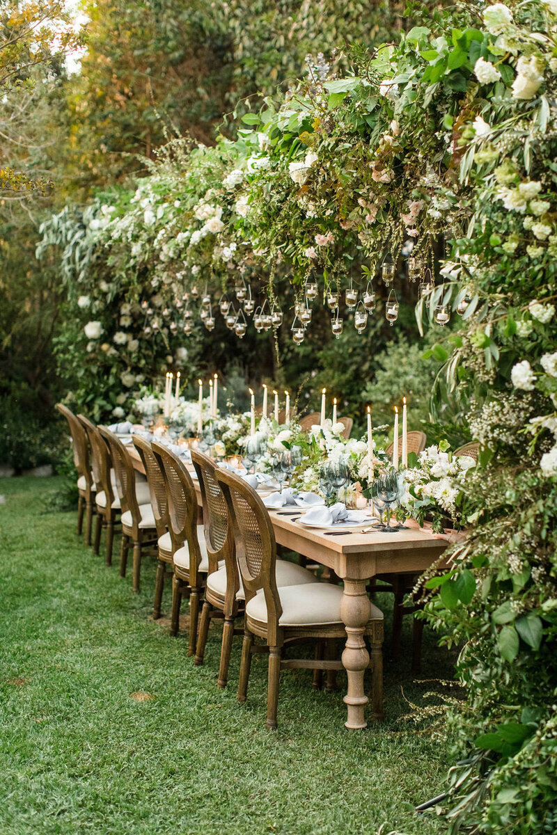 Intimate outdoor wedding reception set up featuring one long grand wooden table, with a huge floral arch spanning the length of the table.