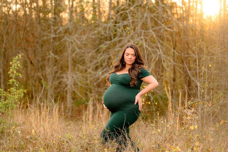 Dark-haired expectant mom poses with one hand on the back of her hip and the other hand on her belly and a hunter green maternity dress that's tight fitting to show off her pregnant silhouette