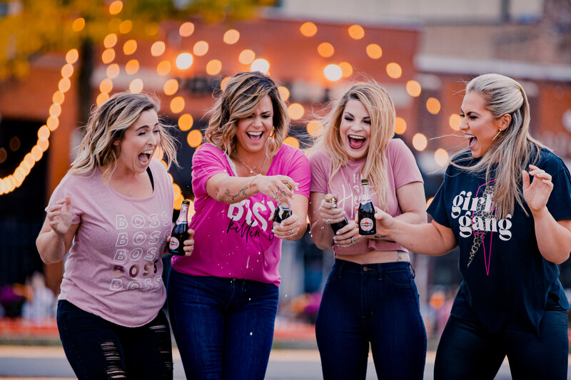 Four women laughing and screaming while popping mini champagne bottles and the champagne is spraying. There are twinkle lights behind them.