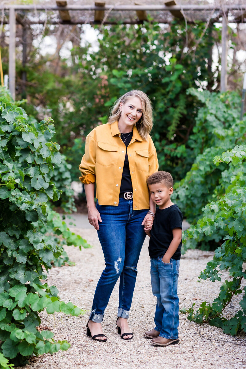 Mother and son mommy and me outdoor mini session.  Mom is wearing a yellow jacket and black shirt with jeans and son is wearing a black shirt with blue jeans.  Both are looking at the camera while holding hands and standing in a vineyard.