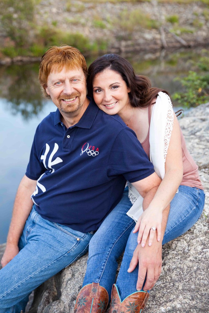 A smiling couple seated on rocks with a serene lake in the background; the man is wearing a dark polo shirt with USA Olympics logo, and the woman is in a white top with lace details.