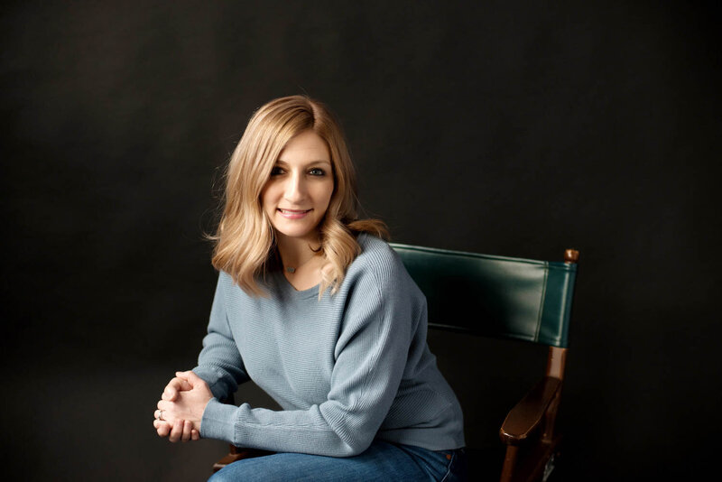 professional head shot of blond woman sitting in chair wearing grey sweater.