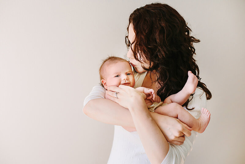 A mom kisses her baby on the cheek while the baby smiles and looks at the camera in Daniele Rose Photography's studio