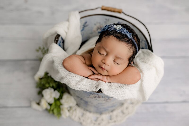 close-up image of newborn baby girl posed in a bucket on a white layer with a blue bonnet headband
