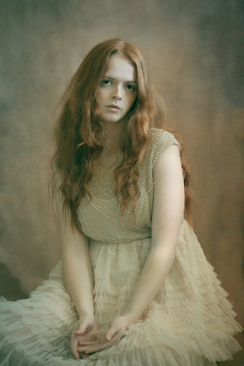 A girl with red hair sits in a tulle dress with her hands clasped