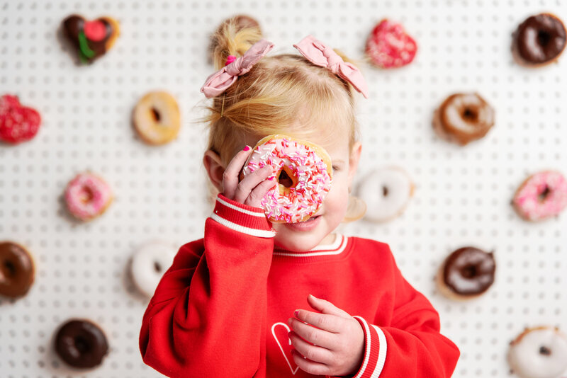 st-louis-mini-sessions-little-girl-holding-donut-to-face-in-front-of-valentines-day-donut-wall