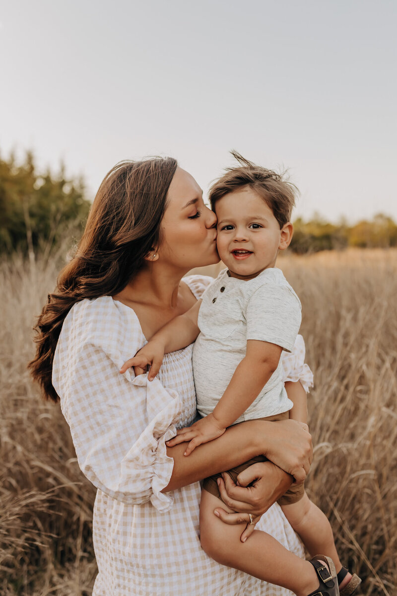 A mom in a cream flowy dress holds her toddler son wearing a cream button up. They are walking through a field for a photoshoot by San Antonio photographer.