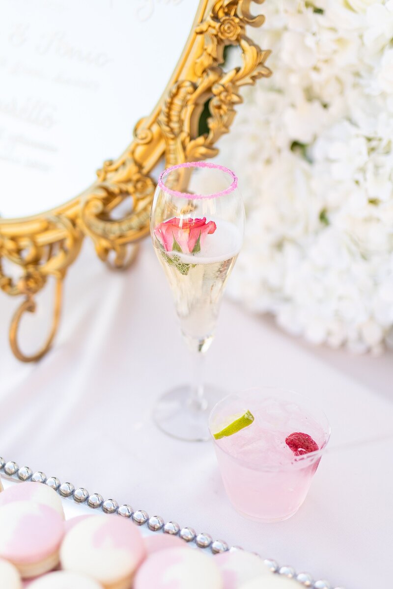 Rose champagne drink and raspberry and lime cocktail with pink macaron in front of white flower wall and golden mirror.